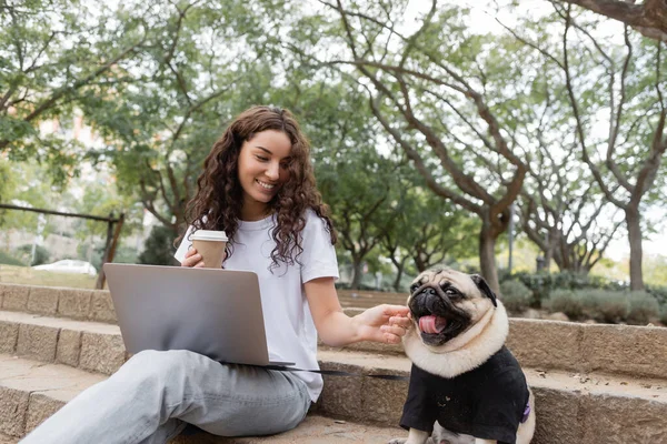 Smiling and curly freelancer in casual clothes holding coffee to go near laptop and petting pug dog while sitting on stairs in blurred park in Barcelona, Spain — Stock Photo