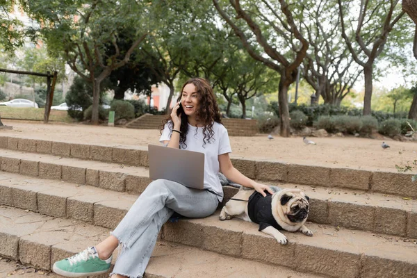 Cheerful brunette freelancer in casual clothes talking on smartphone near laptop and petting pug dog on stairs while spending time in blurred park in Barcelona, Spain — Stock Photo