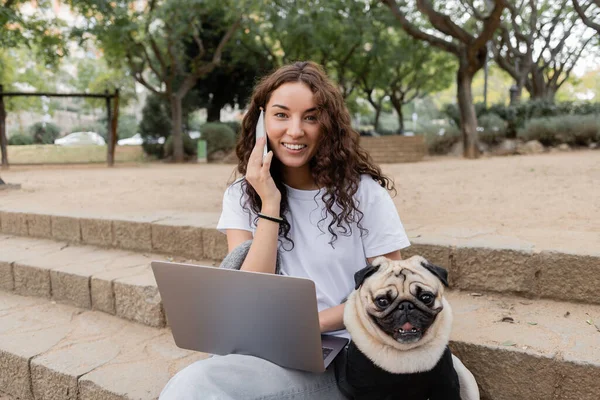 Carefree young curly woman in casual clothes looking at camera while talking on cellphone near laptop and pug dog sitting on stairs in blurred park in Barcelona, Spain — Stock Photo