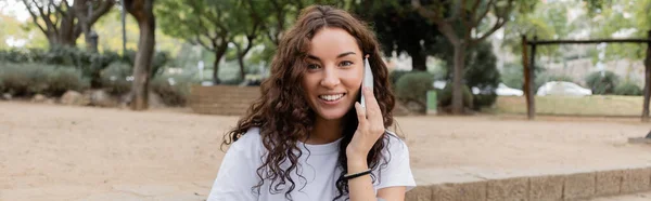 Portrait de jeune femme bouclée en t-shirt blanc souriant à la caméra tout en parlant sur smartphone et en passant du temps dans un parc flou de jour à Barcelone, Espagne, bannière — Photo de stock