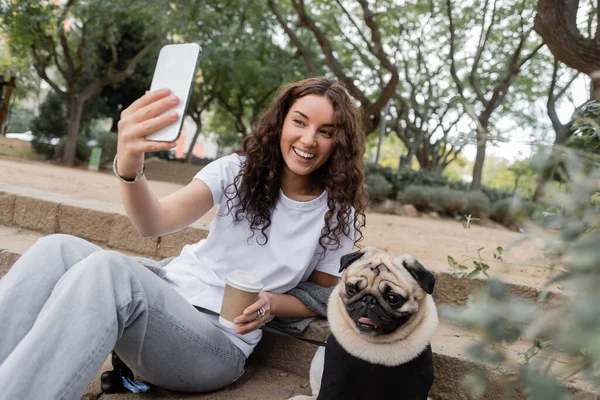 Carefree young and curly woman in casual clothes having video call on cellphone and holding coffee to go while sitting near pug dog on stairs in blurred park at daytime in Barcelona, Spain — Stock Photo