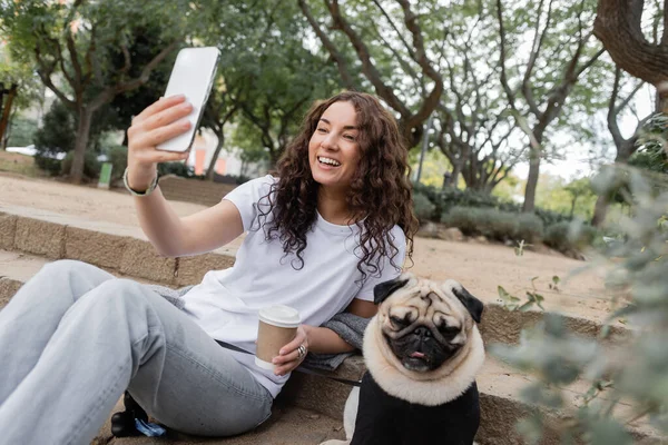 Happy young woman in casual clothes having video chat on smartphone and holding coffee to go while sitting near pug dog on stairs in blurred park at daytime in Barcelona, Spain — Stock Photo