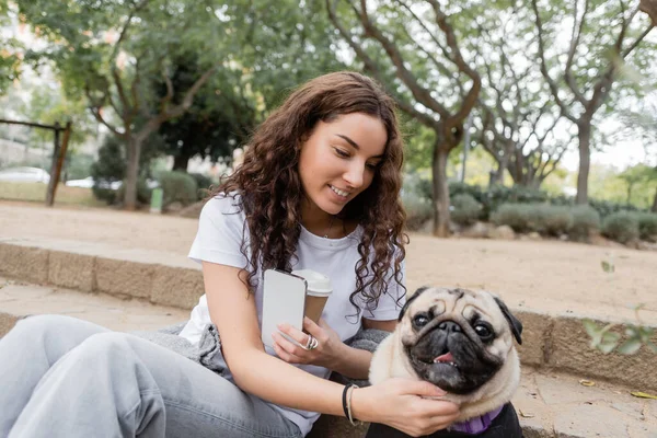 Pretty and curly young woman in casual clothes holding smartphone and coffee to go while petting pug dog on stairs and spending time in blurred park in Barcelona, Spain — Stock Photo