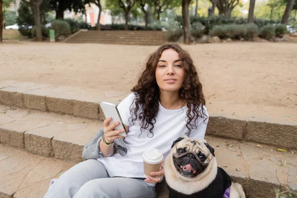 Smiling young curly woman in casual clothes using mobile phone and holding coffee to go while looking at camera near pug dog sitting on stairs in blurred park in Barcelona, Spain — Stock Photo