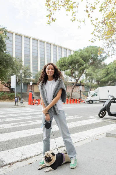 Беззаботная молодая женщина в повседневной одежде смотрит в камеру и держит поводок, стоя рядом с собакой-мопсом возле здания на размытой городской улице днем в Барселоне, Испания — стоковое фото