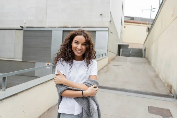 Portrait of smiling curly and pretty woman in white t-shirt and sweater looking at camera while standing near blurred industrial building at background on city street in Barcelona, Spain — Stock Photo