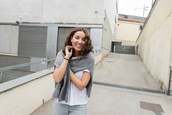 Веселая молодая и кудрявая женщина в белой футболке трогает теплый свитер и смотрит в камеру, стоя рядом с размытым промышленным зданием на городской улице в Барселоне, Испания — стоковое фото