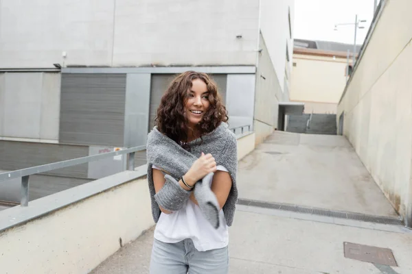 Улыбающаяся молодая и кудрявая женщина в повседневной одежде, держащая теплый серый свитер и глядя в сторону, стоя рядом с размытым промышленным зданием на городской улице Барселоны, Испания — стоковое фото