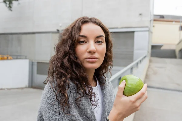 Portrait d'une jeune femme bouclée en pull chaud tenant une pomme verte fraîche et regardant une caméra sur une rue urbaine floue à Barcelone, en Espagne, le jour, bâtiment industriel — Photo de stock