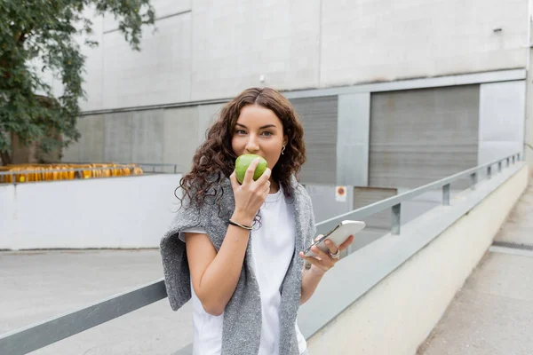 Femme frisée et brune avec pull regardant la caméra tout en mangeant de la pomme verte fraîche et en utilisant un smartphone près d'un bâtiment industriel flou dans la rue urbaine de Barcelone, Espagne — Photo de stock