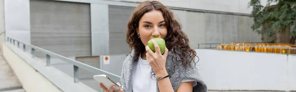 Jeune femme brune avec un pull chaud sur les épaules manger pomme verte mûre et tenant smartphone tout en regardant la caméra dans la rue urbaine de Barcelone, Espagne, bannière, bâtiment industriel — Photo de stock