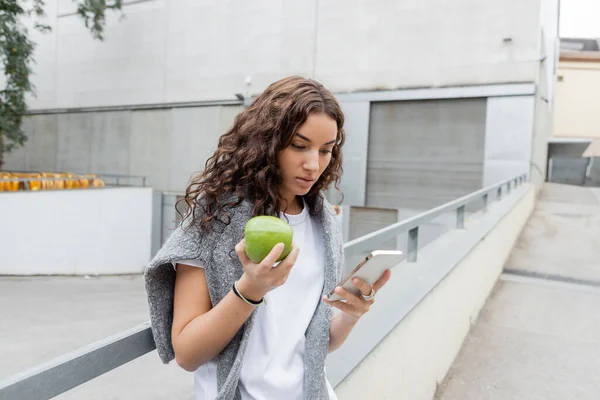 Mujer joven y bonita con suéter cálido en hombros usando celular y sosteniendo manzana fresca mientras está de pie en la calle urbana borrosa con edificio industrial en Barcelona, España - foto de stock
