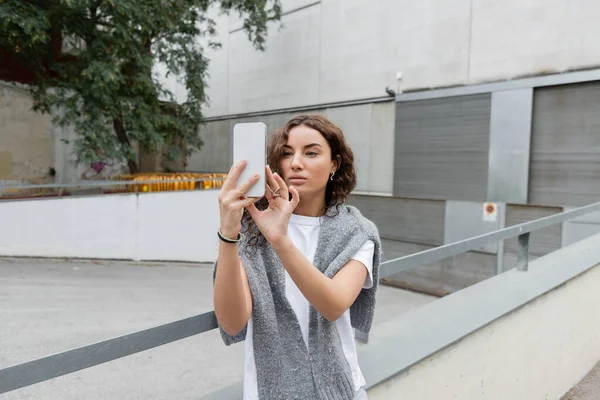 Pretty brunette woman with warm sweater on shoulders using mobile phone while standing on urban street with blurred industrial building at background in Barcelona, Spain — Stock Photo