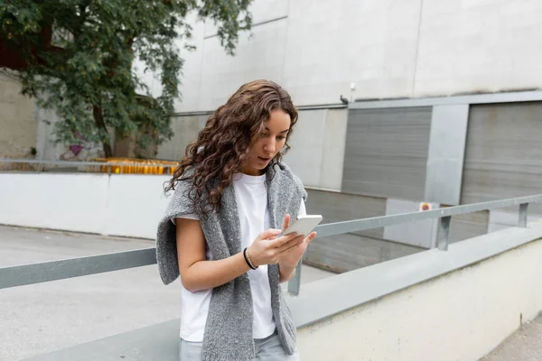 Young curly woman in warm sweater on shoulders using smartphone while standing on city street with buildings at background at daytime in Barcelona, Spain — Stock Photo