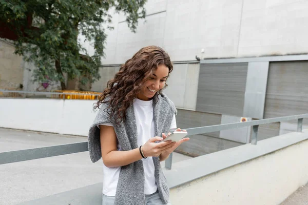 Радостная молодая кудрявая женщина в сером свитере на плечах с помощью смартфона стоит на городской улице с размытыми зданиями днем в Барселоне, Испания — стоковое фото