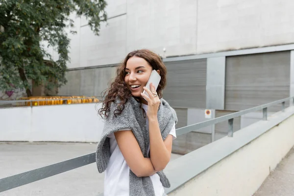 Cheerful and young curly woman in t-shirt and warm sweater talking on smartphone and looking away while standing near blurred buildings on urban street in Barcelona, Spain, green tree — Stock Photo