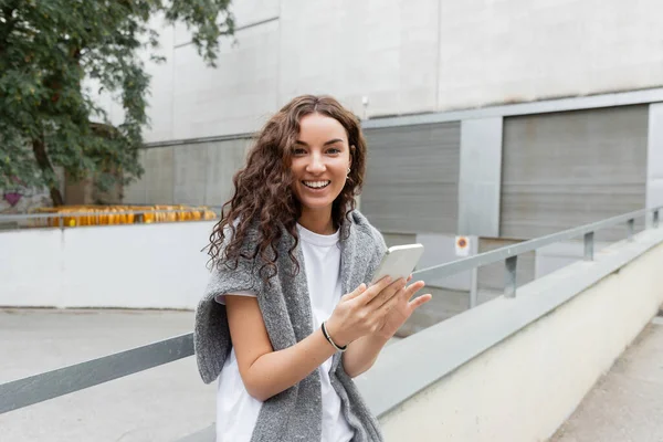 Portrait of positive and curly young woman in t-shirt and sweater looking at camera and using mobile phone while standing on city street at daytime in Barcelona, Spain, green tree — Stock Photo