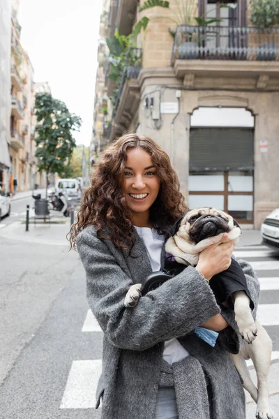Fröhliche junge Frau in warmer Jacke, die in die Kamera blickt und einen Mops auf Händen hält, während sie tagsüber in der Nähe eines verschwommenen Gebäudes auf der städtischen Straße in Barcelona steht, Spanien — Stockfoto