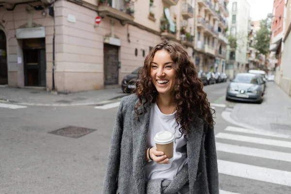 Femme positive jeune et bouclée en veste décontractée tenant du café dans une tasse en papier et regardant loin sur la rue floue de la ville avec des voitures et des bâtiments pendant la journée à Barcelone, Espagne — Photo de stock