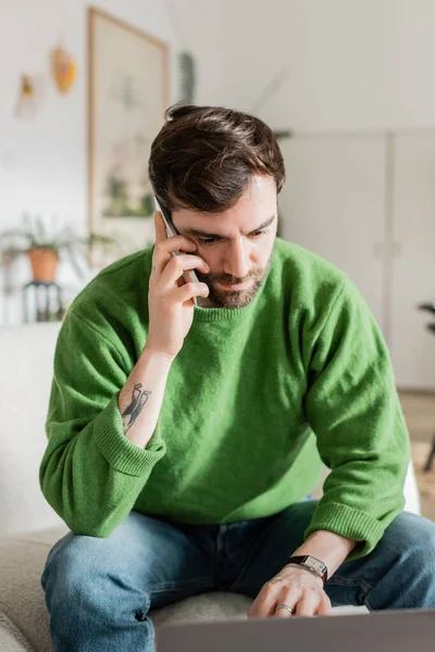 Bearded and tattooed freelancer in casual clothes talking on smartphone and using blurred laptop — Stock Photo