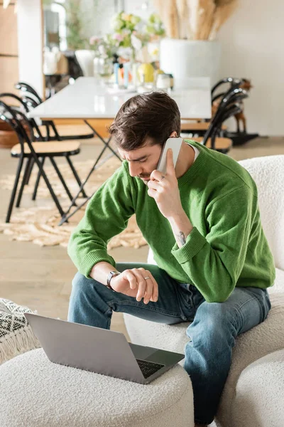Man in green jumper and jeans looking at wristwatch and talking on smartphone while working on laptop — Stock Photo