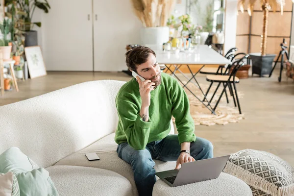 Unternehmer in lässiger Kleidung schaut weg, telefoniert mit dem Smartphone und sitzt auf der Couch neben dem Laptop — Stockfoto