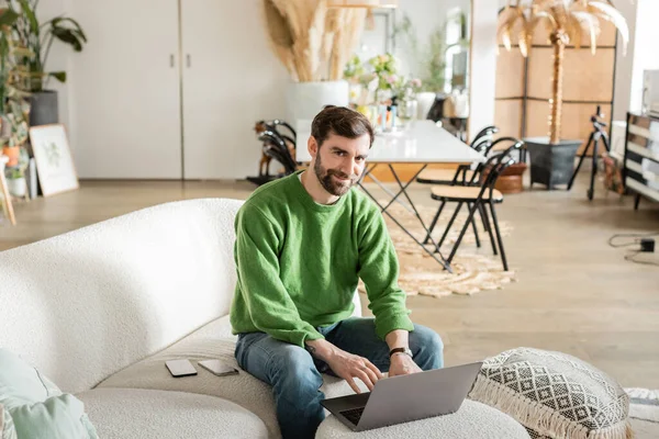 Smiling bearded tattooed freelancer in green jumper and jeans looking at camera while using laptop — Stock Photo