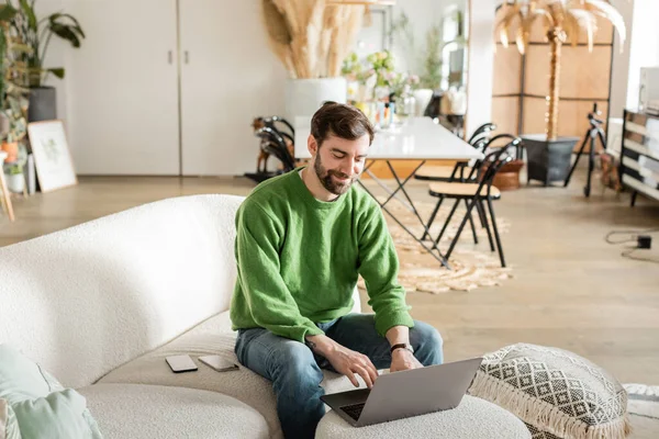 Cheerful bearded freelancer in jumper and jeans working on laptop while sitting near smartphones — Stock Photo