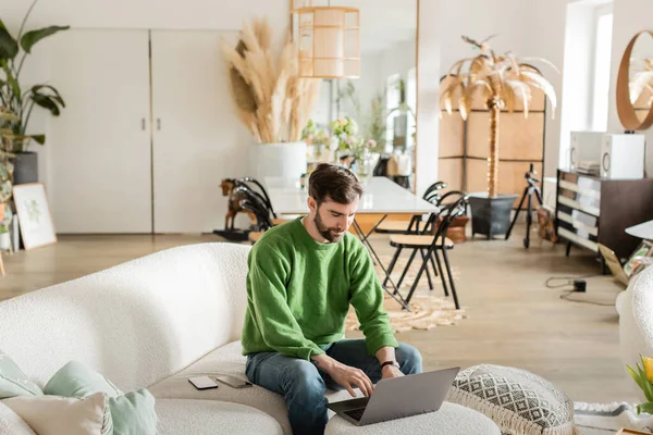 Bearded tattooed freelancer in jumper and jeans using laptop while working and sitting on couch — Stock Photo