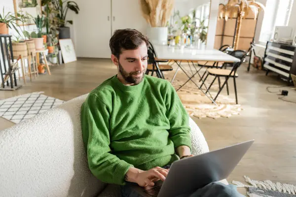 Bärtige und brünette Freiberuflerin in grünem Pullover mit Laptop bei der Arbeit zu Hause — Stockfoto