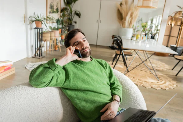 Freelancer barbudo y morena en jersey verde y jeans hablando en smartphone en salón - foto de stock
