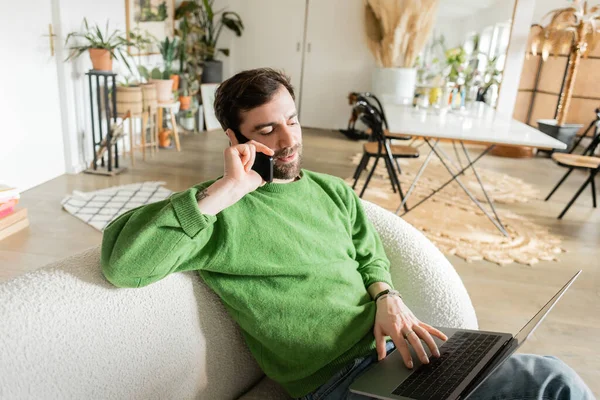 Bearded freelancer in green jumper and jeans talking on smartphone while using laptop at home — Stock Photo