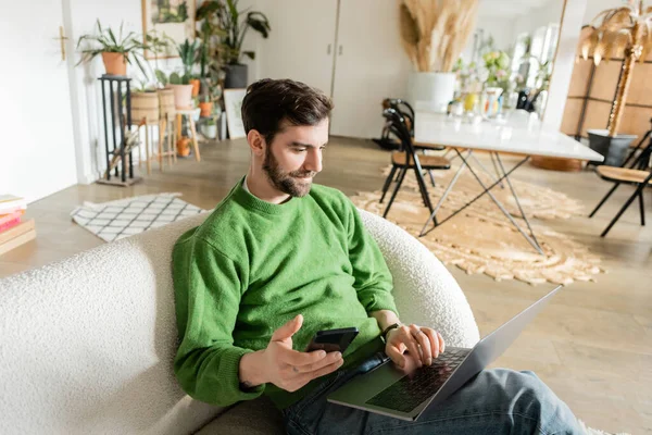 Улыбающийся бородатый фрилансер в зеленом свитере и джинсах с помощью ноутбука и держа смартфон дома — стоковое фото