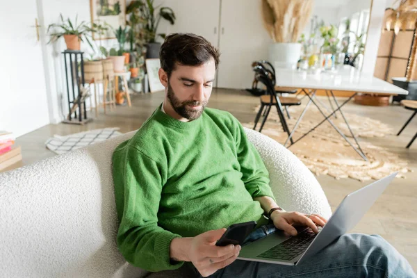 Freelancer barbudo en jersey verde y jeans usando smartphone mientras trabaja en laptop, trabajo remoto - foto de stock