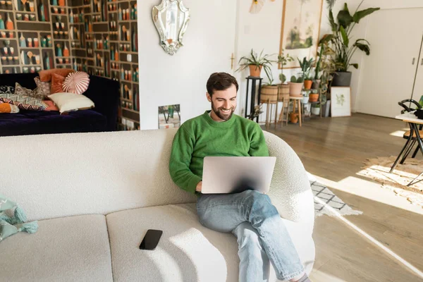 Alegre freelancer barbudo en jersey verde y jeans usando laptop cerca de smartphone, trabajo remoto - foto de stock