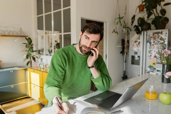 Freelance barbu en pull vert parler sur smartphone et écrire sur ordinateur portable dans la cuisine — Photo de stock