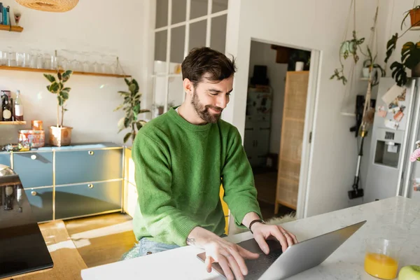 Positive and bearded freelancer in green jumper using laptop while working at home — Stock Photo