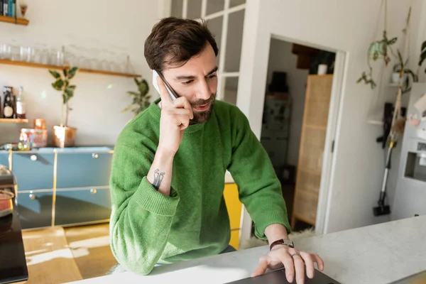 Bearded and tattooed freelancer in jumper talking on smartphone while working on laptop at home — Stock Photo
