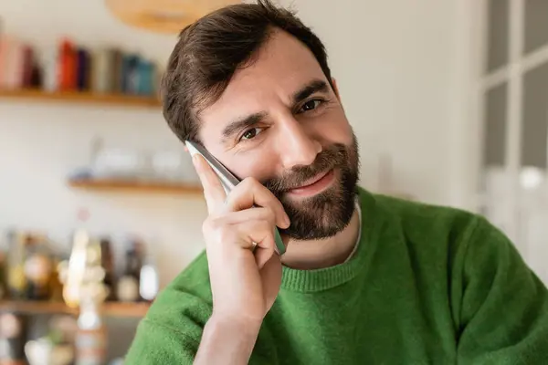 Porträt eines fröhlichen brünetten und bärtigen Mannes im grünen Pullover, der zu Hause mit dem Smartphone spricht — Stockfoto