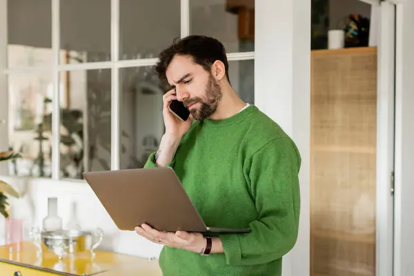 Freelancer enfocado en jumper verde que sostiene el portátil y habla en el teléfono inteligente en casa - foto de stock