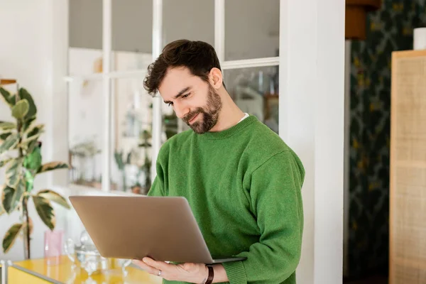 Positive Brünette und bärtige Unternehmerin in grünem Pullover schaut während der Arbeit zu Hause auf den Laptop — Stockfoto