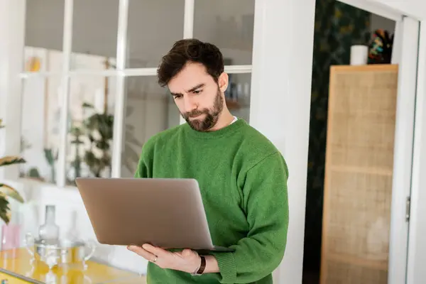 Freelancer barbudo enfocado en jersey verde usando portátil mientras trabaja en casa, trabajo remoto - foto de stock