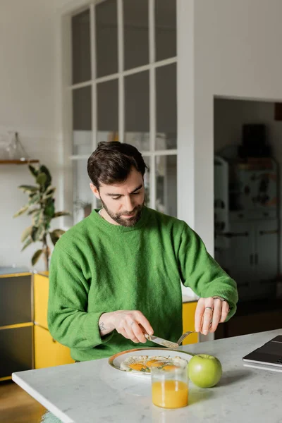 Uomo barbuto in maglione verde con posate vicino alla colazione in cucina moderna a casa — Foto stock