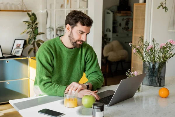 Barbudo y morena hombre en jersey verde utilizando el ordenador portátil cerca de teléfono inteligente y desayuno en la cocina - foto de stock