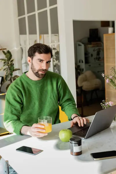 Freelancer barbudo sosteniendo jugo de naranja fresco y el uso de ordenador portátil en la cocina en casa - foto de stock