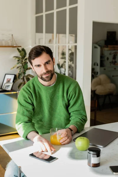 Brünette und bärtige Mann in grünem Pullover hält ein Glas Orangensaft und arbeitet zu Hause — Stockfoto