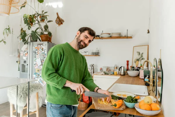 Uomo barbuto sorridente in maglione e jeans che taglia pepe fresco mentre prepara la colazione in cucina — Foto stock