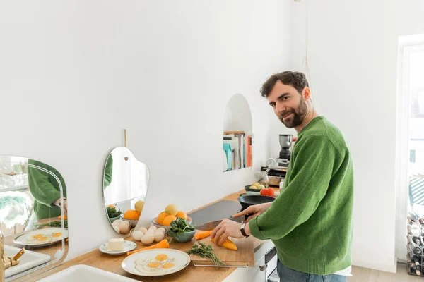 Happy man in green jumper cutting fresh pepper while cooking breakfast in modern kitchen — Stock Photo