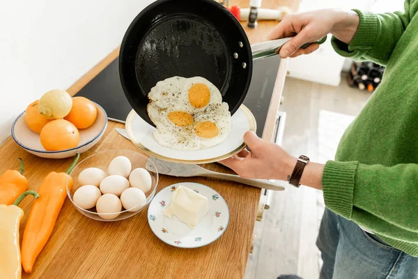 Vista recortada del hombre sosteniendo la sartén con huevos fritos cerca del plato mientras cocina el desayuno por la mañana - foto de stock