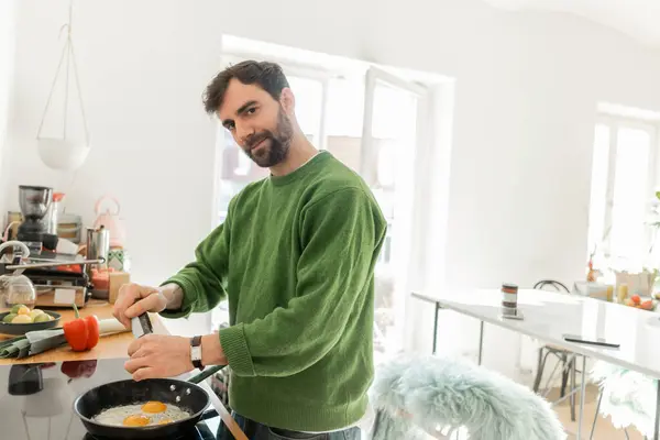 Homem empanado em saltador verde olhando para a câmera enquanto cozinha ovos fritos para o café da manhã — Fotografia de Stock